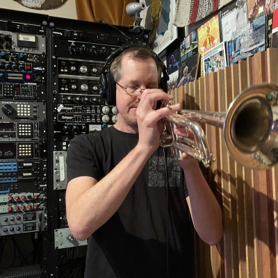 A photo of Erik Jekabson playing a trumpet in front of a rack at Wide Hive Records.