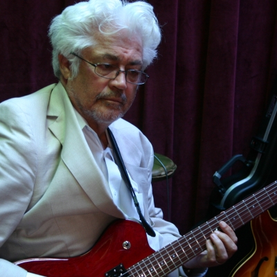 Larry Coryell in a white coat with his eyes closed, playing the guitar. 